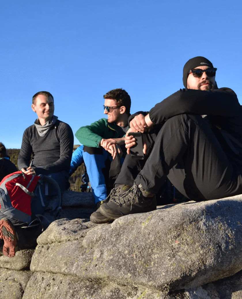 Three people sit on a rocky surface enjoying a sunny day. Two are wearing sunglasses, and all are dressed in outdoor gear. They appear relaxed, surrounded by a clear blue sky.