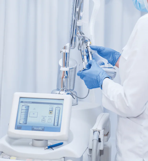 A healthcare professional wearing gloves and a mask uses a medical machine with a control screen. The background is white, and the focus is on the equipment and the person's hands operating it.