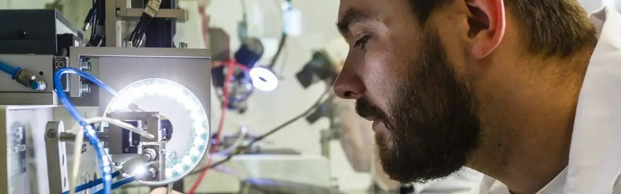 A man with a beard is closely examining a piece of equipment, which is illuminated, in a laboratory setting. He is wearing a white lab coat, and the background is filled with various scientific instruments and blurred lights.