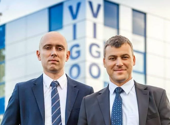 Two men in suits stand in front of a modern building with glass windows. The building has the letters "VIGO" visible in the background. Both men are wearing ties and are looking towards the camera.