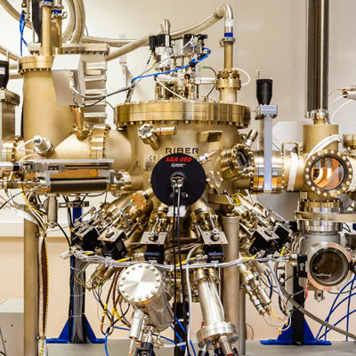 A complex scientific machine with numerous metal pipes, valves, and wires is displayed. A central cylindrical chamber is labeled "Riber" with additional instruments attached. The setup appears to be part of a laboratory experiment or research.
