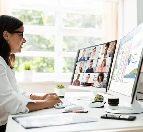 A person sits at a desk working on a computer with two monitors. One screen shows a video conference with nine participants; the other displays charts and data. The room has a bright window with plants nearby.
