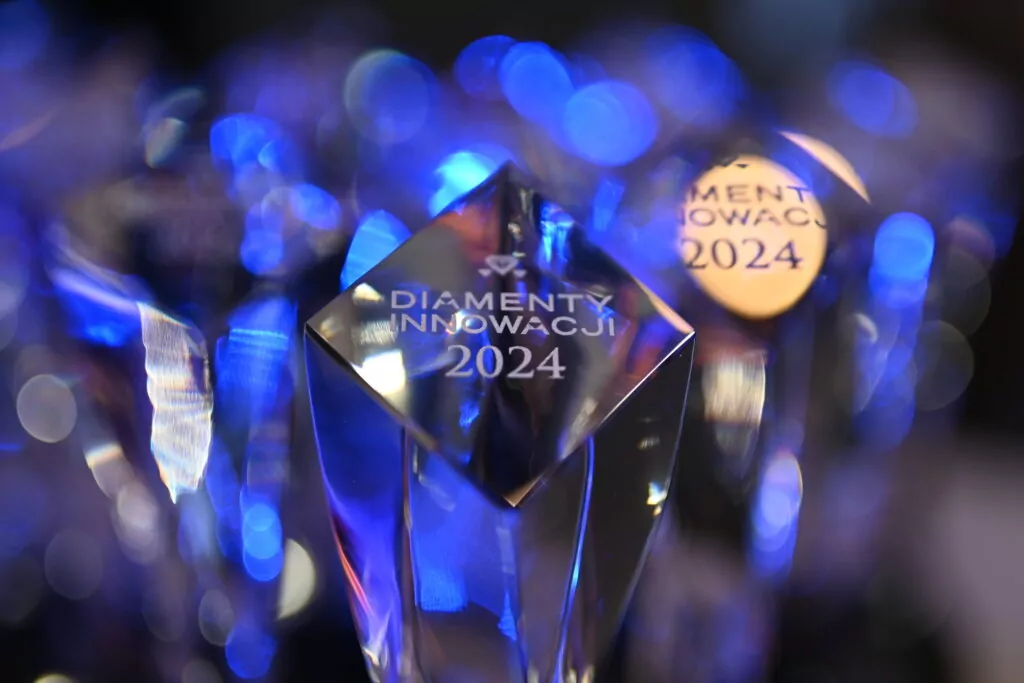 Close-up of a crystal award with the inscription "DIAMENTY INNOWACJI 2024" in white letters. The background consists of similar crystal awards, all reflecting blue light.