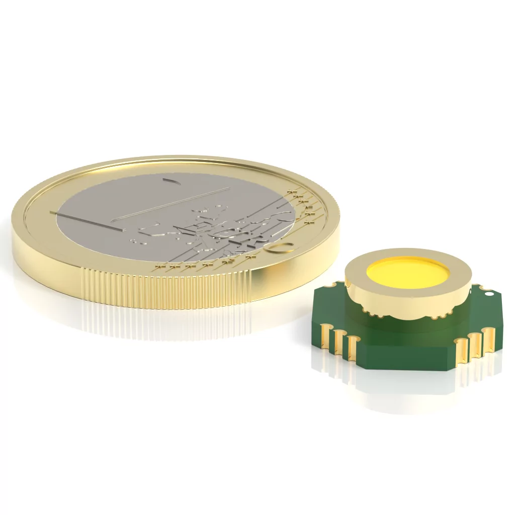 A comparison image showing a gold coin on the left and a small cylindrical electronic component with a green base and yellow top on the right. The component is slightly smaller than the coin and positioned parallel to it.