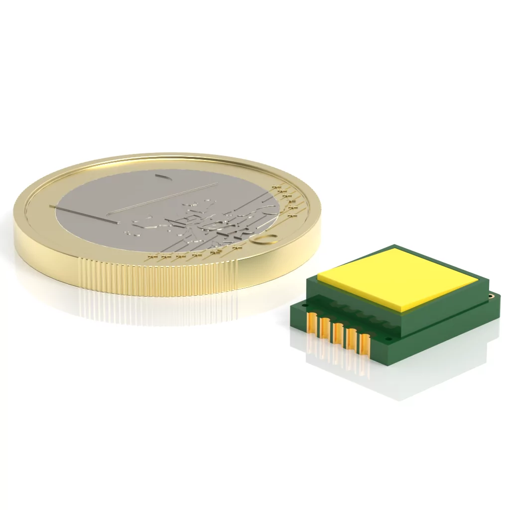 A gold coin and a small green electronic component with gold pins are placed side by side on a reflective surface. The electronic component is rectangular with a flat yellow top.