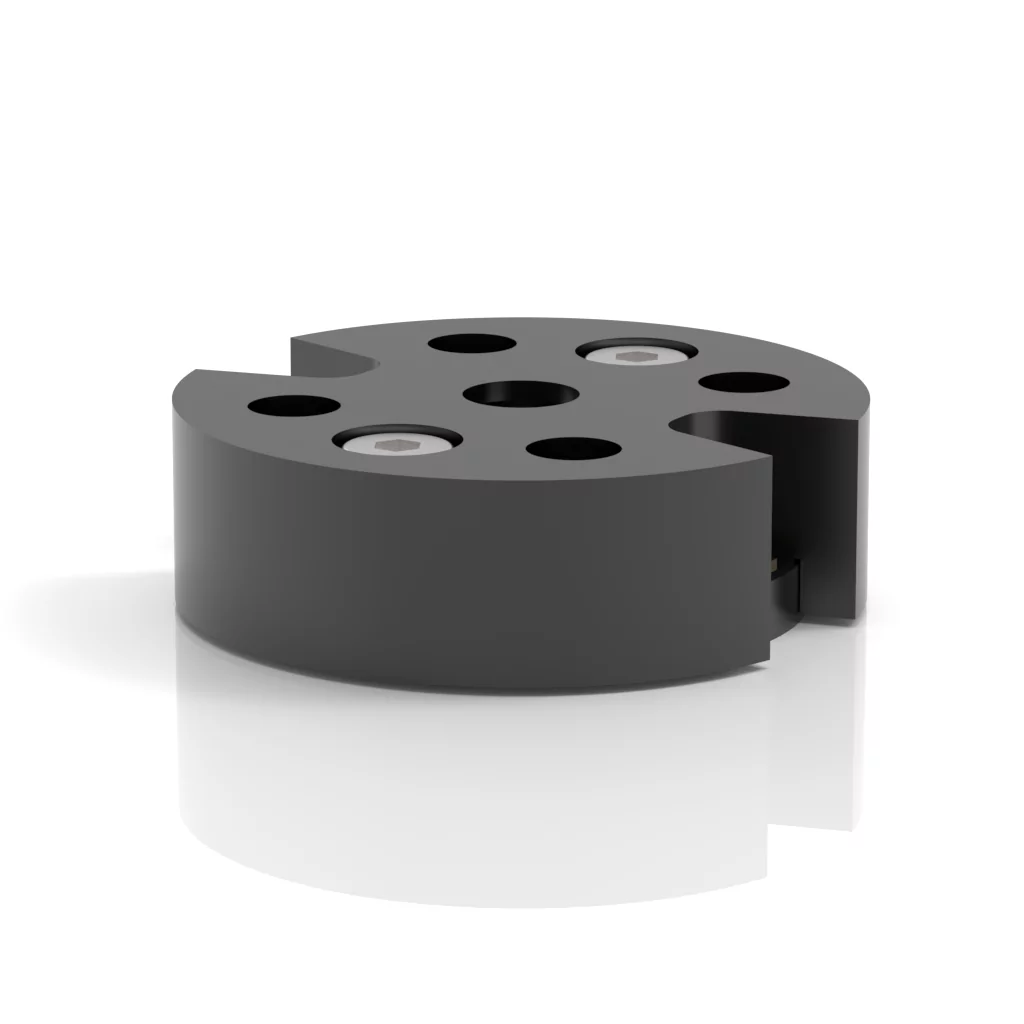A black cylindrical mechanical part with multiple holes of varying sizes, resting on a reflective white surface. The part features a cutout section and appears industrial in design.