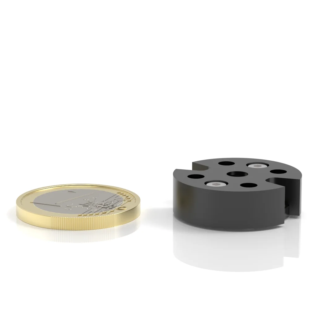 A gold-colored coin lies flat next to a small, round, black cylindrical object with holes and screws, both on a white surface with reflections.
