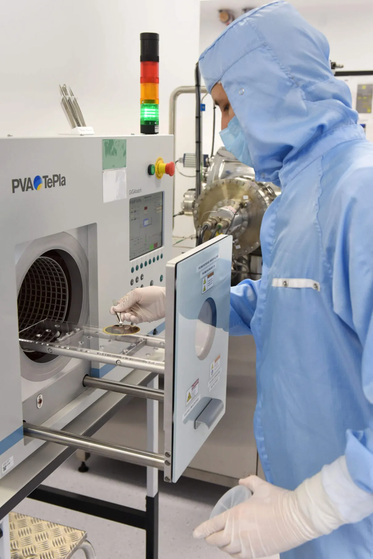A person in a blue cleanroom suit and gloves operates a PVA TePla machine, placing items inside. The environment appears to be a laboratory with scientific equipment visible in the background.