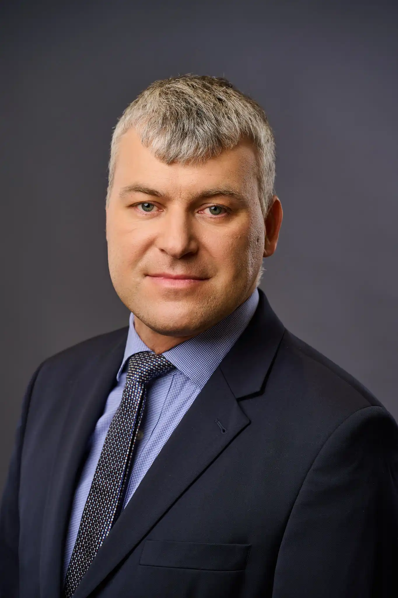 A man in a navy suit, light blue shirt, and patterned tie poses for a professional headshot against a plain dark background. He has short, light gray hair and a slight smile.