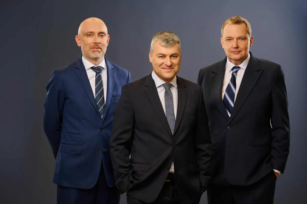 Three men in suits pose together against a plain, dark background. They are standing close, facing the camera, and all are dressed formally in business attire with ties.