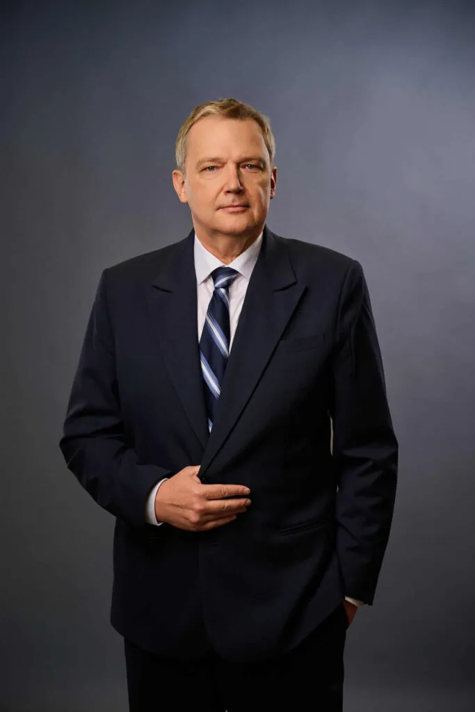 A middle-aged man in a dark suit and striped tie stands against a gray studio background, with one hand holding his jacket and the other in his pocket, looking confidently at the camera.