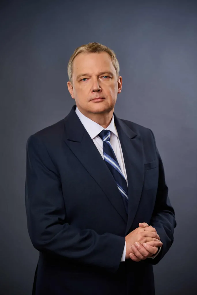 A middle-aged man in a dark suit and striped tie stands against a plain, dark background, looking seriously at the camera with his hands clasped in front of him.
