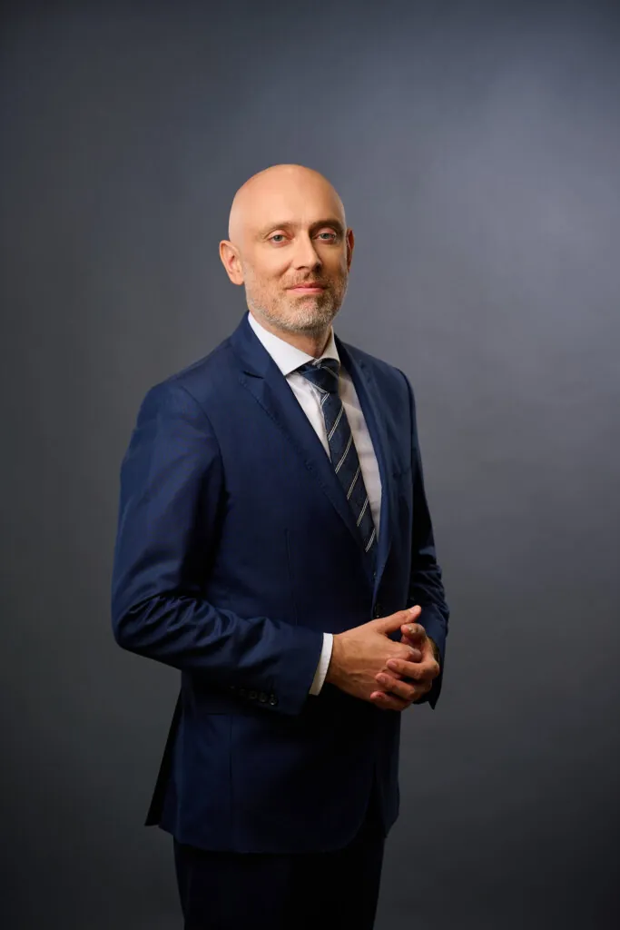 A bald man with a trimmed beard wearing a blue suit, white shirt, and striped tie stands against a plain gray background, posing with his hands clasped and a slight smile.