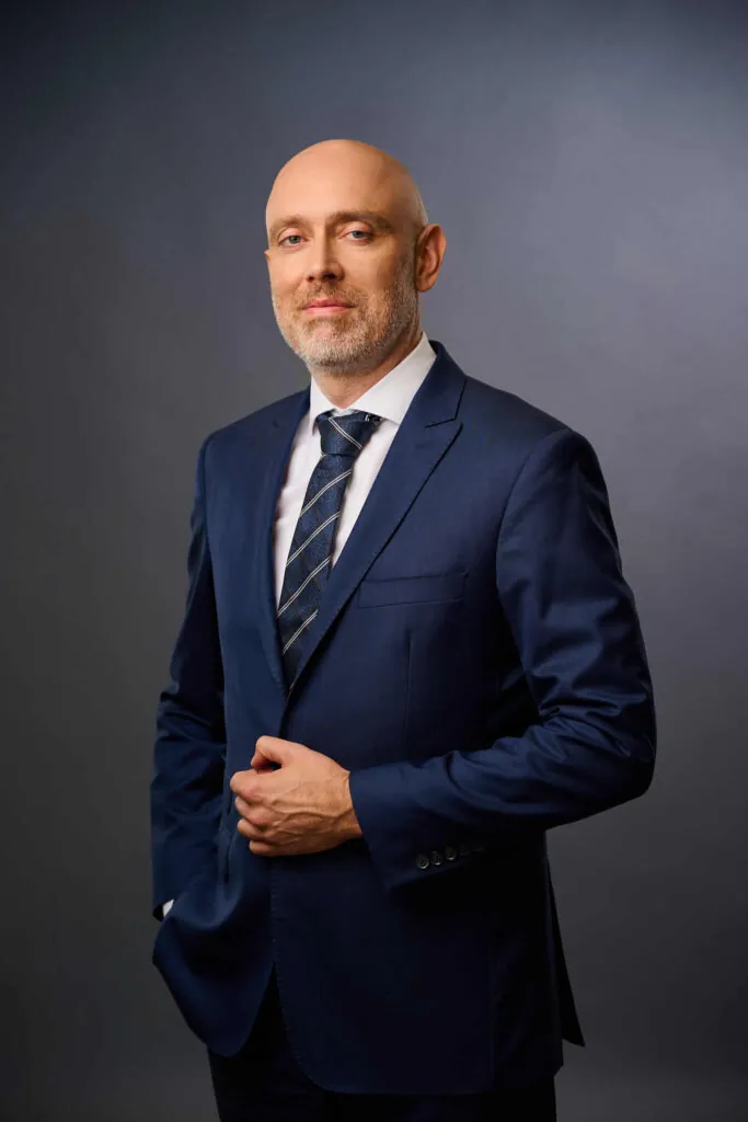 A confident bald man with a trimmed beard wearing a navy blue suit, white shirt, and striped tie stands against a plain dark gray background, posing with one hand on his suit jacket.