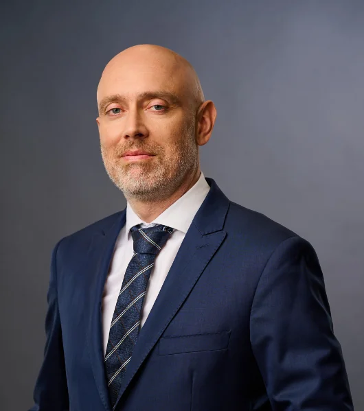 A bald man with a trimmed beard wearing a navy blue suit, white dress shirt, and striped tie stands against a plain gray background, looking confidently at the camera.
