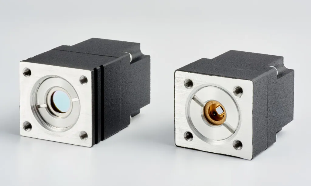 Two black and silver electronic components with square metal faces and circular centers, each featuring four corner screws, are placed side by side on a plain, light background.