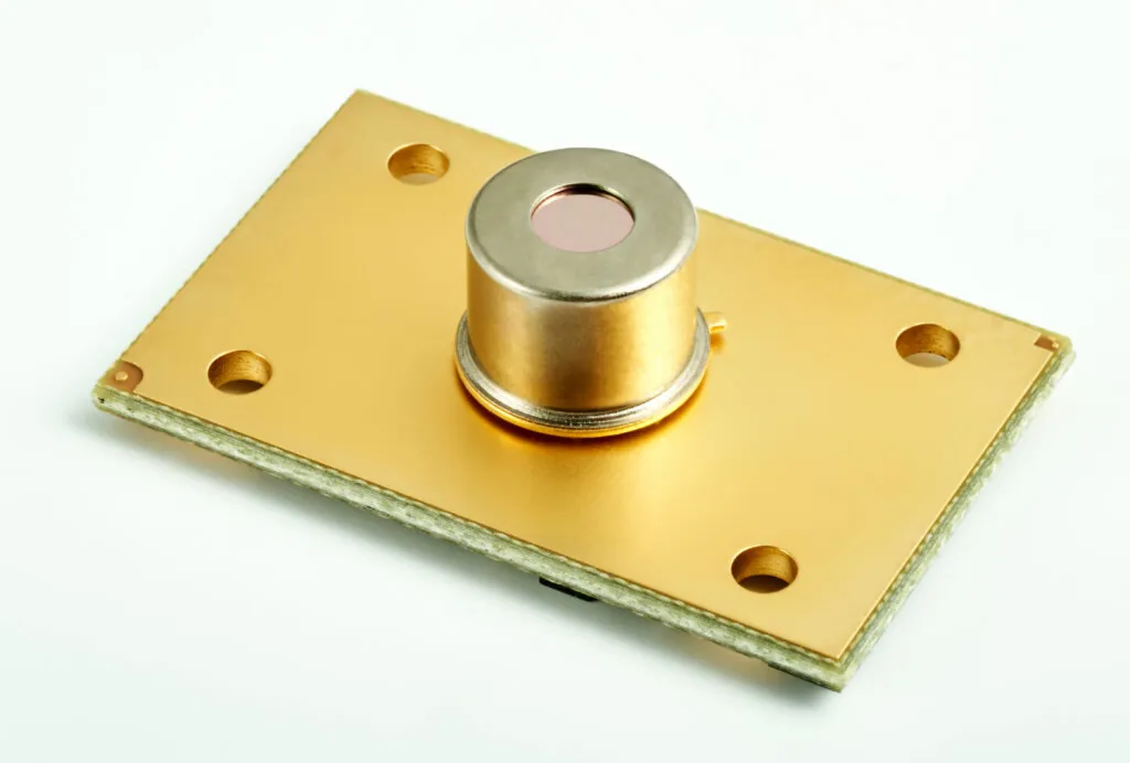 A gold-colored electronic sensor with a cylindrical component mounted on a rectangular base plate featuring four corner holes, placed on a white background.