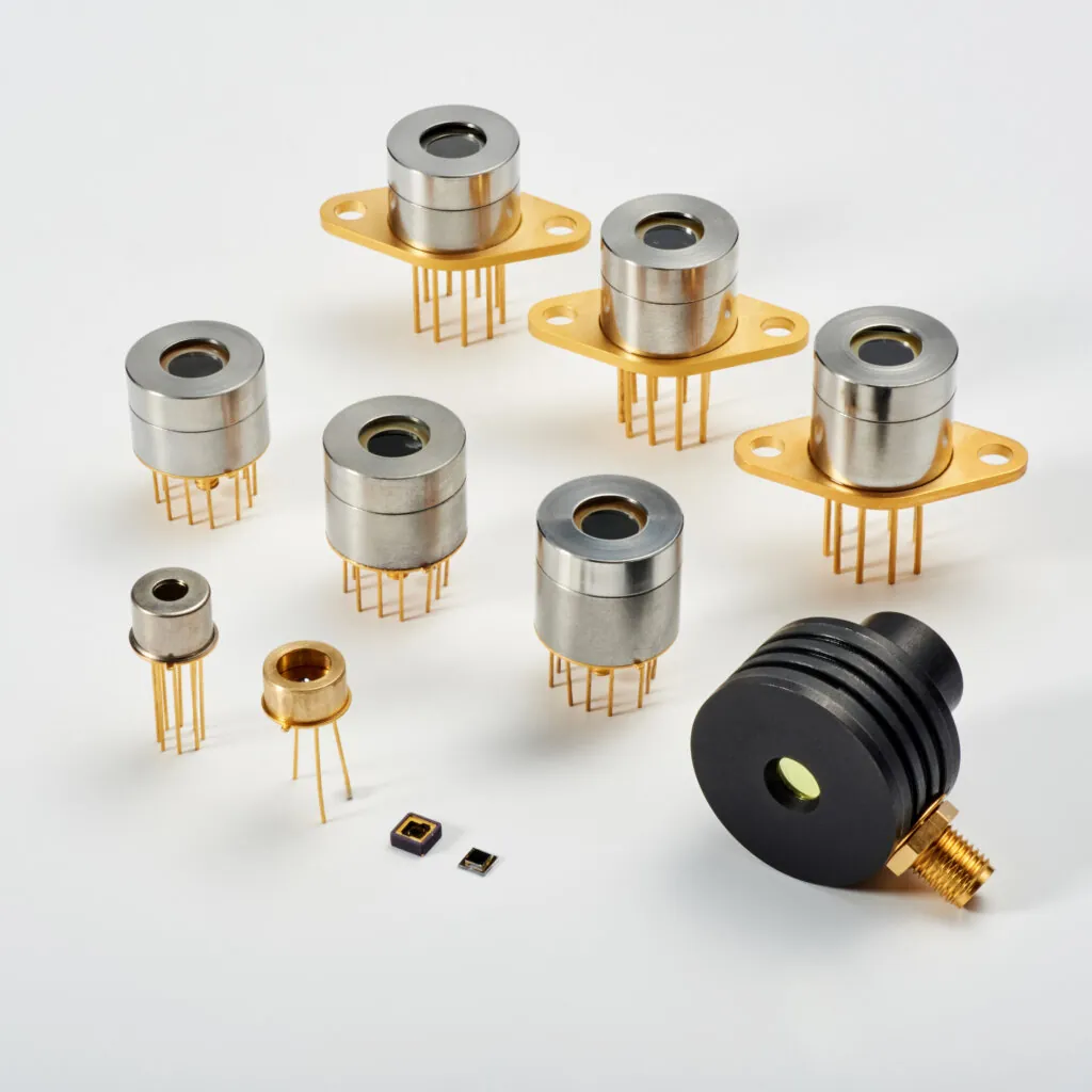 A collection of various metal and gold electronic components with pins, arranged on a white surface. Some components have a cylindrical shape, and others are flat and square.