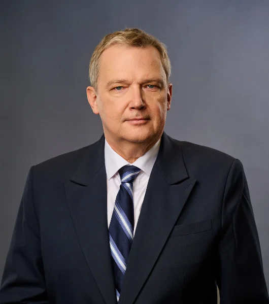A middle-aged man in a dark suit, light blue shirt, and striped tie stands against a plain gray background, looking directly at the camera with a neutral expression.