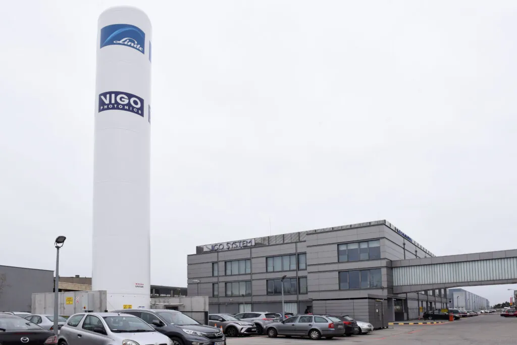 A large white industrial tower labeled Linde and Vigo Photonics stands beside a modern gray building complex with several parked cars and a skybridge on a cloudy day.