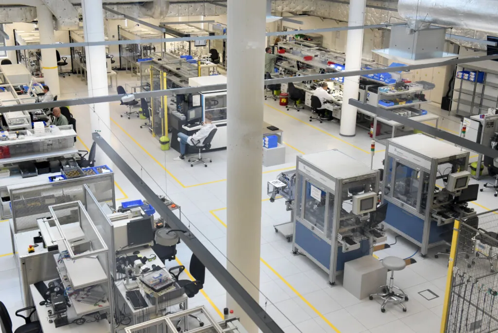 A clean, modern factory floor with automated machinery, workstations, and equipment arranged in organized rows. Few workers are visible, and the area is brightly lit with white walls and floors.