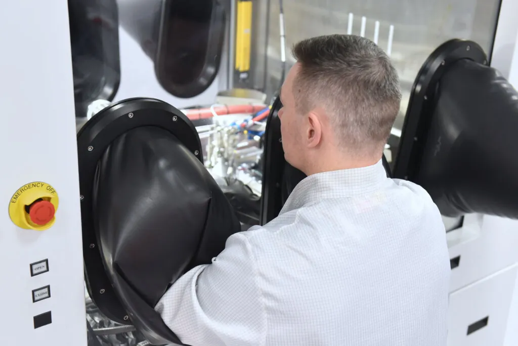 A person wearing a lab coat operates equipment inside a containment glove box, using black gloves attached to the enclosure. An emergency stop button is visible on the machine.