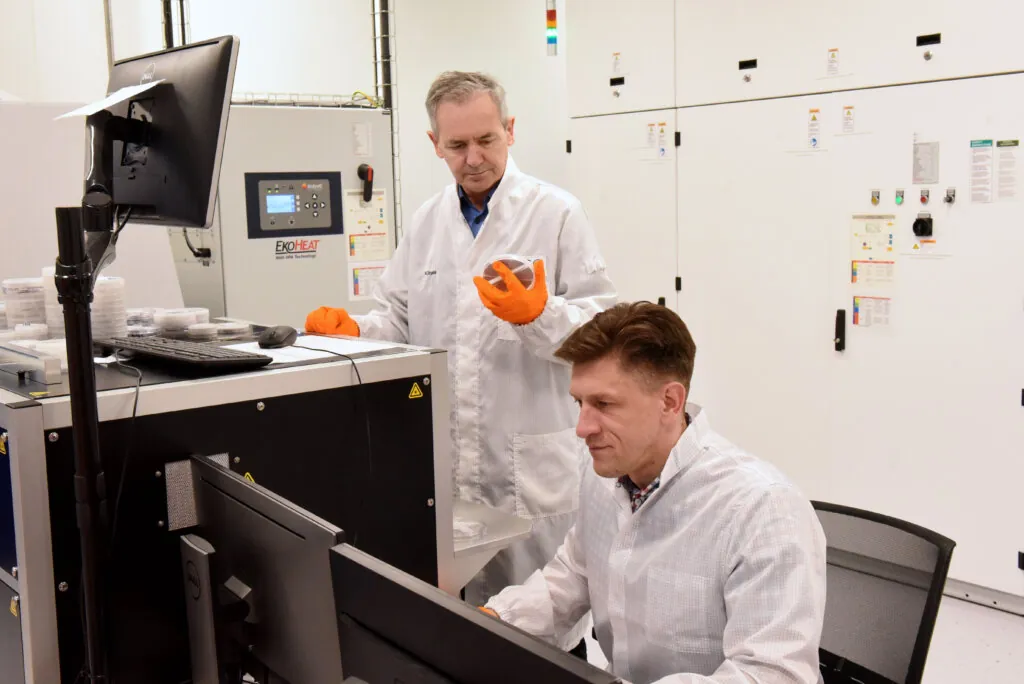 Two men in lab coats work in a laboratory. One is seated at a computer, focused on the screen, while the other stands nearby, wearing orange gloves and gesturing as he observes the workstation. Laboratory equipment surrounds them.