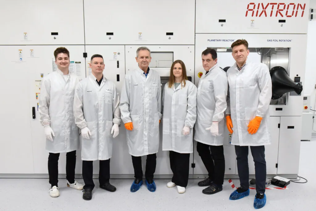 Six people wearing white lab coats and gloves stand in a row in front of large laboratory equipment labeled AIXTRON. They are posing for a group photo in a clean, modern laboratory setting.