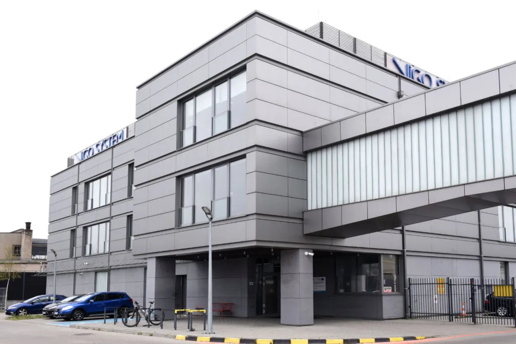 A modern gray office building with large windows, an elevated glass walkway, and a parking lot with several cars and bicycles. The building has a sign on the roof with blue lettering.