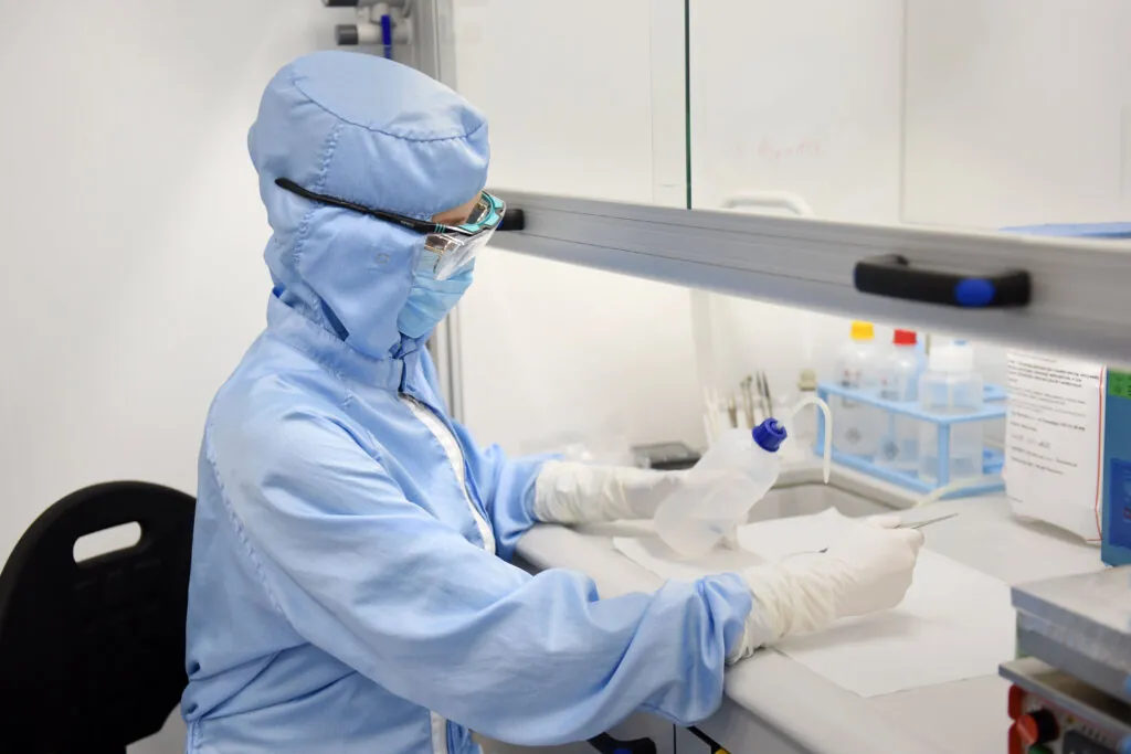 A scientist in full protective gear, including a mask, gloves, and goggles, works in a laboratory cleanroom, holding a plastic bottle and using lab equipment on a sterile work surface.