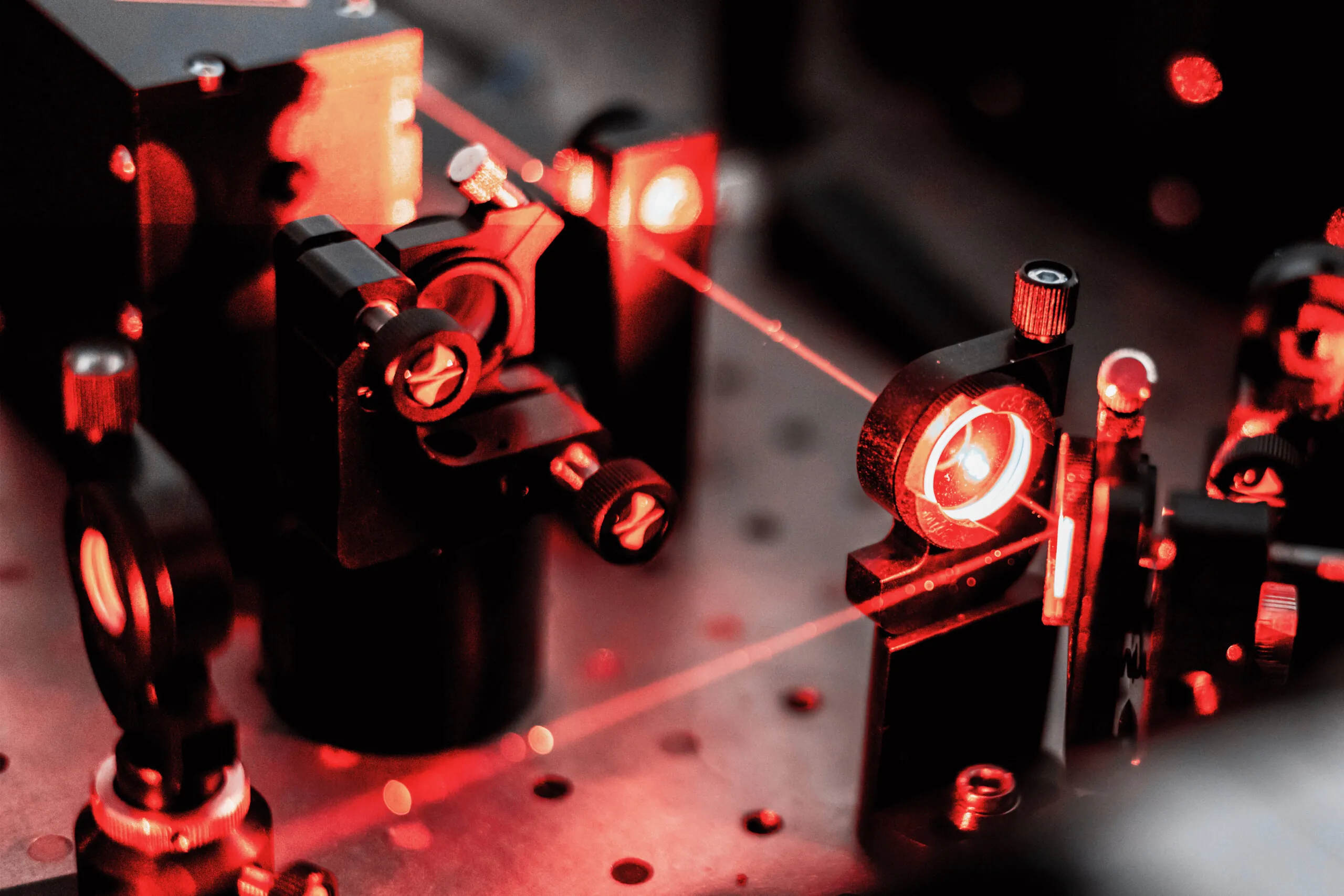 A close-up of a scientific optical setup with red laser beams passing through and reflecting off various lenses and mirrors mounted on a perforated metal surface.